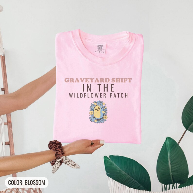 Blossom Comfort Colors t-shirt printed with the words Graveyard Shift In The Wildflower Patch and a drawing of a cute chubby ghost wearing a flower crown and holding a single violet, folded flat lay