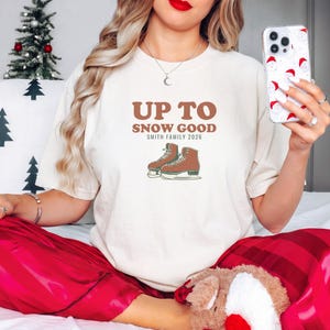 Ivory Comfort Colors t-shirt printed with the words Up To Snow Good Smith Family 2026 and a drawing of a vintage metal clamp-on ice skate with red laces, worn by a woman