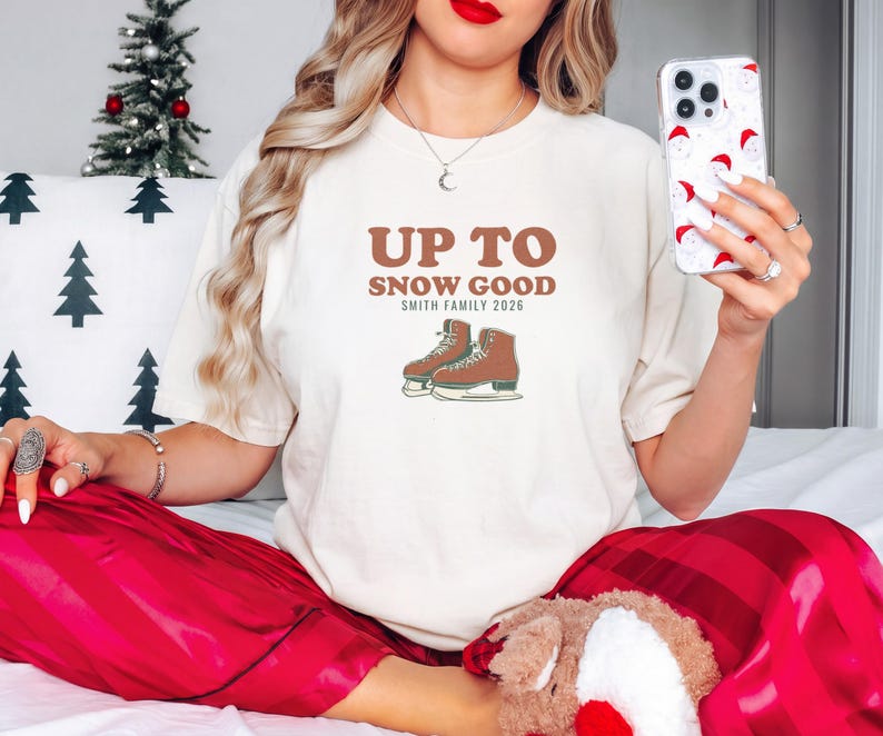 Ivory Comfort Colors t-shirt printed with the words Up To Snow Good Smith Family 2026 and a drawing of a vintage metal clamp-on ice skate with red laces, worn by a woman