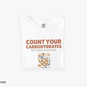 White Comfort Colors t-shirt printed with the words Count Your Carbohydrates Not Your Blessings and a drawing of a golden-brown braided pretzel with coarse salt grains, folded flat lay