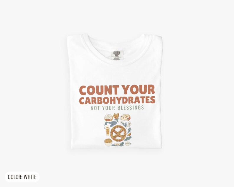 White Comfort Colors t-shirt printed with the words Count Your Carbohydrates Not Your Blessings and a drawing of a golden-brown braided pretzel with coarse salt grains, folded flat lay