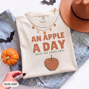 Ivory Comfort Colors t-shirt printed with the words An Apple A Day Keeps The Chaos Away and a drawing of a shiny red apple with a single green leaf and a tiny worm peeking out, worn by a woman
