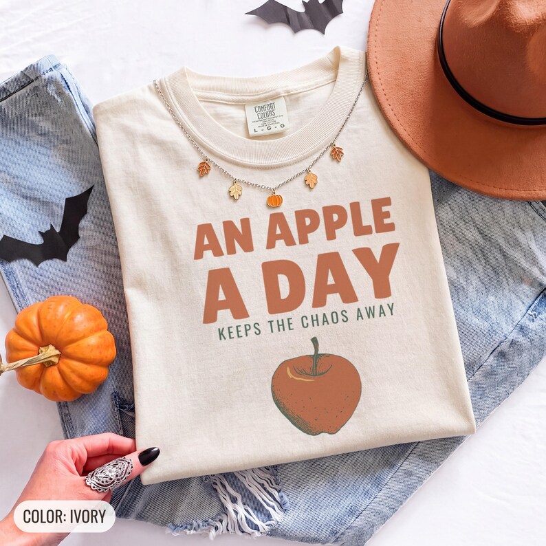 Ivory Comfort Colors t-shirt printed with the words An Apple A Day Keeps The Chaos Away and a drawing of a shiny red apple with a single green leaf and a tiny worm peeking out, worn by a woman