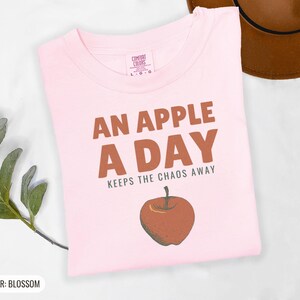 Blossom Comfort Colors t-shirt printed with the words An Apple A Day Keeps The Chaos Away and a drawing of a shiny red apple with a single green leaf and a tiny worm peeking out, folded flat lay