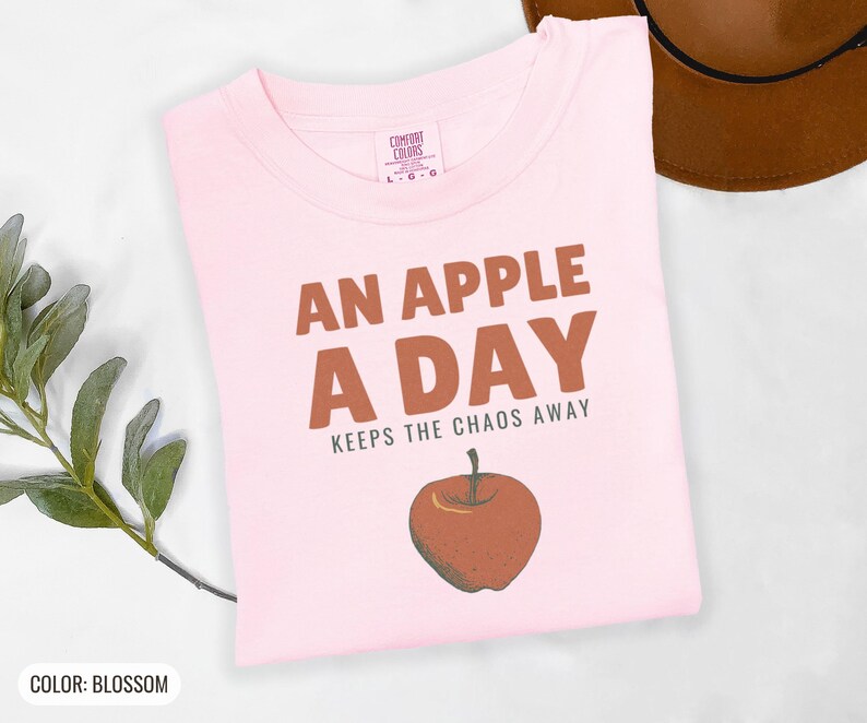 Blossom Comfort Colors t-shirt printed with the words An Apple A Day Keeps The Chaos Away and a drawing of a shiny red apple with a single green leaf and a tiny worm peeking out, folded flat lay