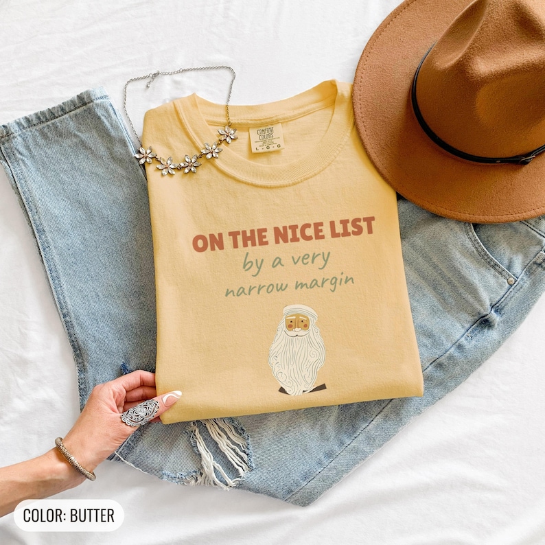 Butter Comfort Colors t-shirt printed with the words On The Nice List By A Very Narrow Margin and a drawing of a red santa hat beside a candy cane, folded flat lay
