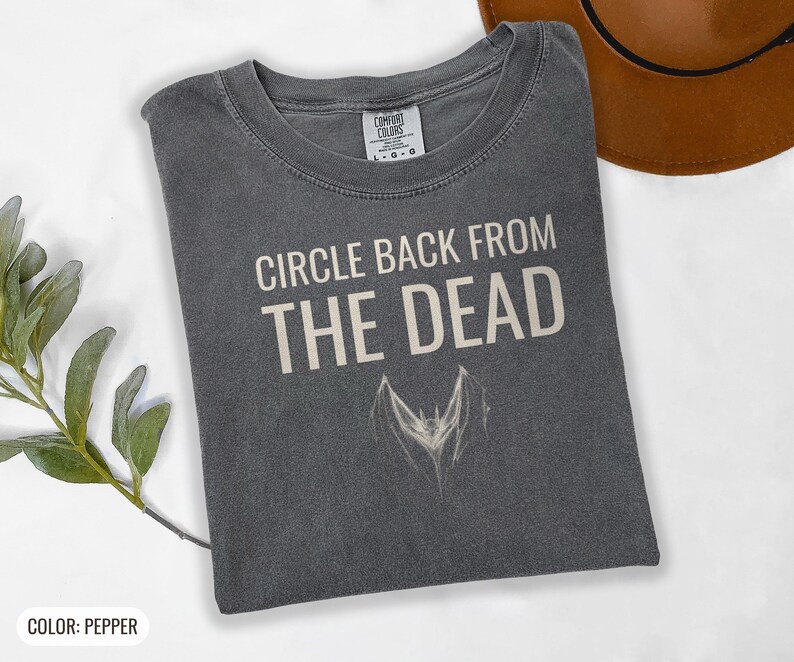 Pepper Comfort Colors t-shirt printed with the words Circle Back From The Dead and a drawing of a vintage silver paperclip bent into the shape of a bat, folded flat lay