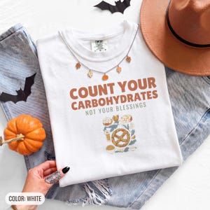 White Comfort Colors t-shirt printed with the words Count Your Carbohydrates Not Your Blessings and a drawing of a golden-brown braided pretzel with coarse salt grains, worn by a woman