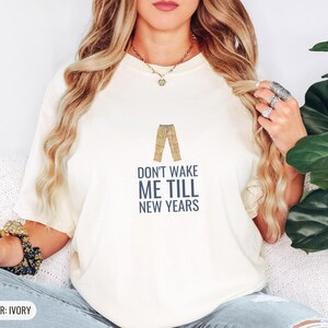 Ivory Comfort Colors t-shirt printed with the words Don'T Wake Me Till New Years and a drawing of a pair of plaid pajama pants folded neatly on a wooden stool, worn by a woman