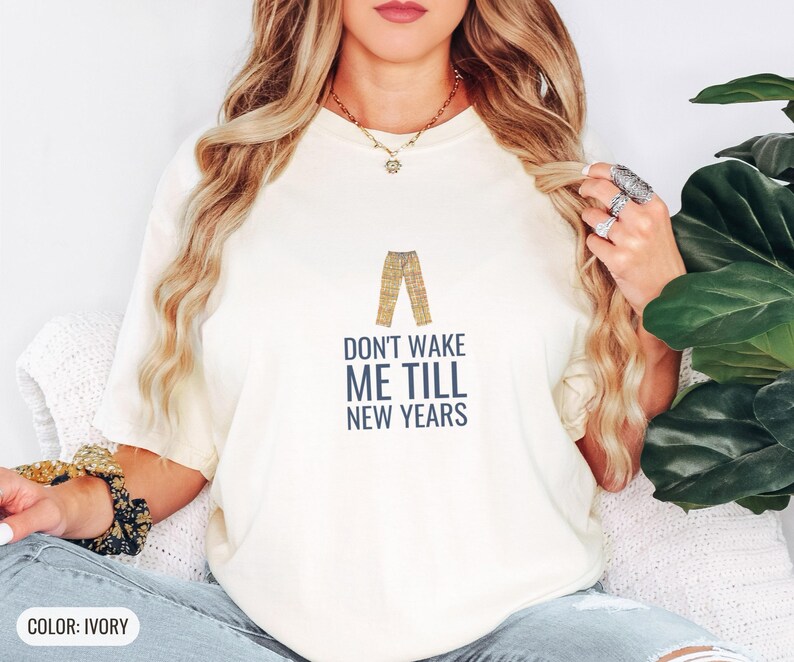 Ivory Comfort Colors t-shirt printed with the words Don'T Wake Me Till New Years and a drawing of a pair of plaid pajama pants folded neatly on a wooden stool, worn by a woman