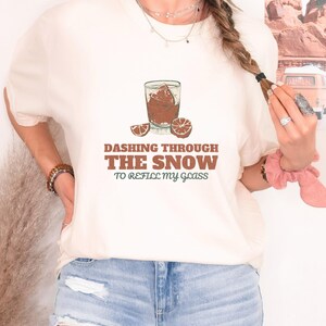 Ivory Comfort Colors t-shirt printed with the words Dashing Through The Snow To Refill My Glass and a drawing of a heavy crystal whiskey tumbler with two floating ice cubes beside a sprig of pine needles, worn by a woman