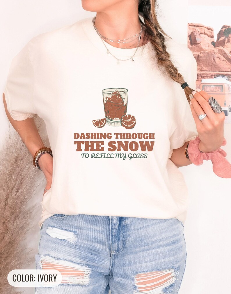 Ivory Comfort Colors t-shirt printed with the words Dashing Through The Snow To Refill My Glass and a drawing of a heavy crystal whiskey tumbler with two floating ice cubes beside a sprig of pine needles, worn by a woman