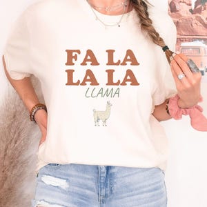 Ivory Comfort Colors t-shirt printed with the words Fa La La La Llama and a drawing of a fluffy llama, worn by a woman