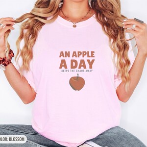 Blossom Comfort Colors t-shirt printed with the words An Apple A Day Keeps The Chaos Away and a drawing of a shiny red apple with a single green leaf and a tiny worm peeking out, worn by a woman