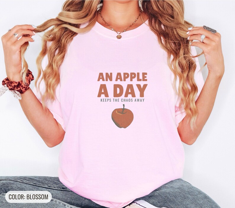 Blossom Comfort Colors t-shirt printed with the words An Apple A Day Keeps The Chaos Away and a drawing of a shiny red apple with a single green leaf and a tiny worm peeking out, worn by a woman
