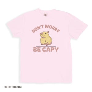 Blossom Comfort Colors t-shirt printed with the words Don'T Worry Be Capy and a drawing of a happy capybara with a small orange citrus slice on its head beside a green lily pad, folded flat lay