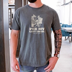 Pepper Comfort Colors t-shirt printed with the words Per My Last Email I Am Deceased and a drawing of a skeleton sitting at an office desk typing on a computer keyboard, worn by a man