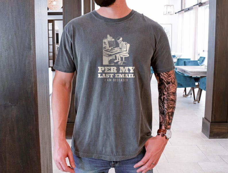 Pepper Comfort Colors t-shirt printed with the words Per My Last Email I Am Deceased and a drawing of a skeleton sitting at an office desk typing on a computer keyboard, worn by a man