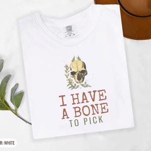White Comfort Colors t-shirt printed with the words I Have A Bone To Pick and a drawing of an anatomical etching of a corvid raven skull entwined with dried wild thistle blossoms, folded flat lay