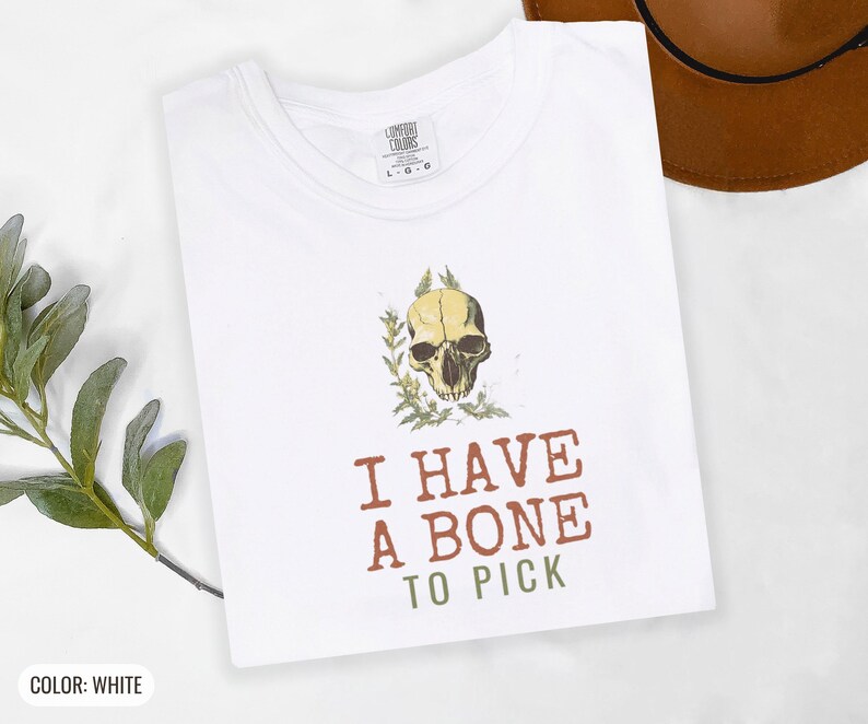 White Comfort Colors t-shirt printed with the words I Have A Bone To Pick and a drawing of an anatomical etching of a corvid raven skull entwined with dried wild thistle blossoms, folded flat lay