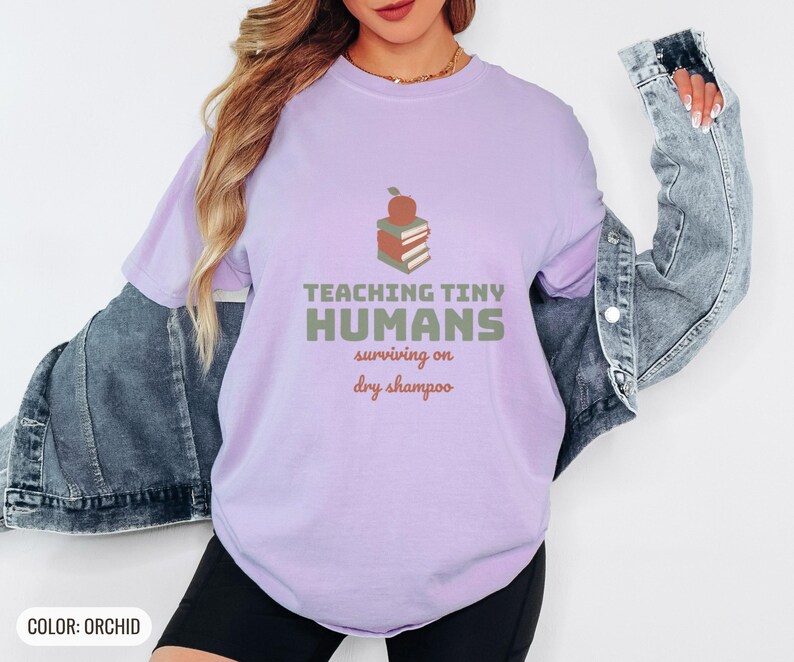 Orchid Comfort Colors t-shirt printed with the words Teaching Tiny Humans Surviving On Dry Shampoo and a drawing of a small neat stack of three closed hardcover books with PLAIN blank solid-colour spines an, worn by a woman