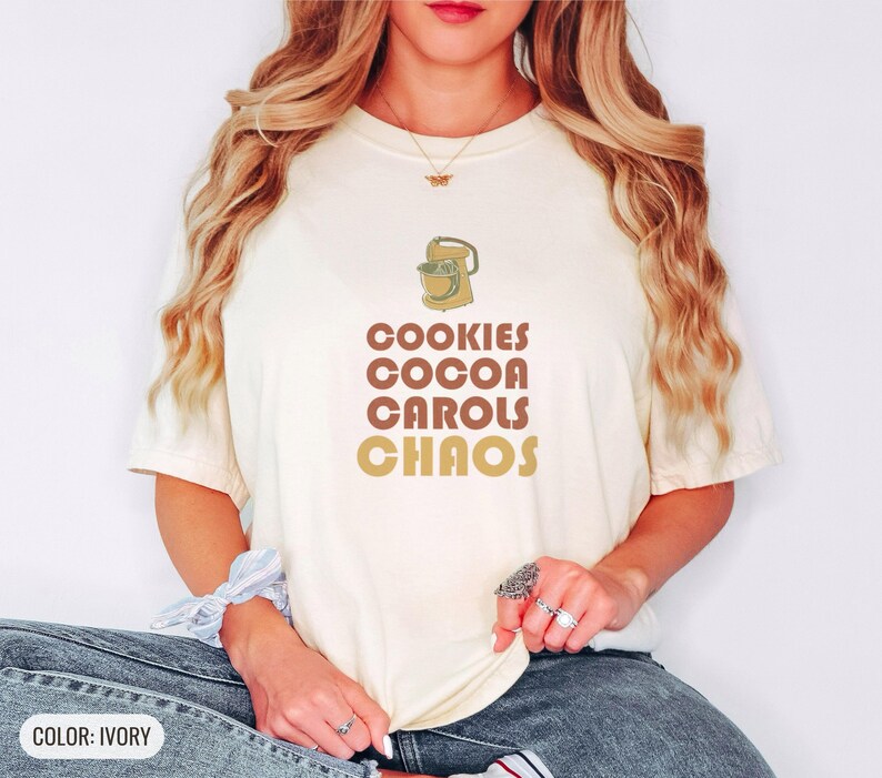 Ivory Comfort Colors t-shirt printed with the words Cookies Cocoa Carols Chaos and a drawing of a retro metal hand mixer with steel beaters, worn by a woman
