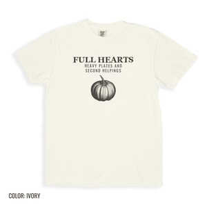 Ivory Comfort Colors t-shirt printed with the words Full Hearts Heavy Plates And Second Helpings and a drawing of a pumpkin surrounded by fall leaves and small acorns, folded flat lay