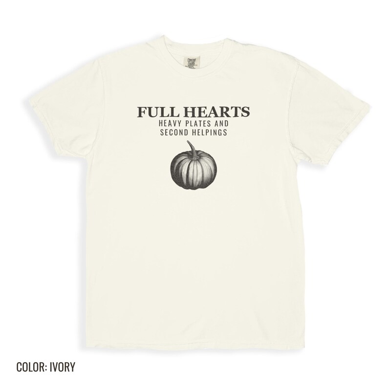 Ivory Comfort Colors t-shirt printed with the words Full Hearts Heavy Plates And Second Helpings and a drawing of a pumpkin surrounded by fall leaves and small acorns, folded flat lay