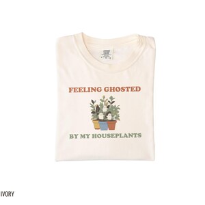 Ivory Comfort Colors t-shirt printed with the words Feeling Ghosted By My Houseplants and a drawing of a compact, self-contained group of three small potted houseplants with tiny, friendly whit, folded flat lay