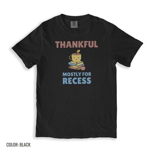 Black Comfort Colors t-shirt printed with the words Thankful Mostly For Recess and a drawing of a stack of retro school books topped with a glossy red apple and a steaming ceramic coffee, folded flat lay
