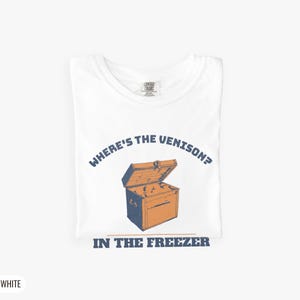 White Comfort Colors t-shirt printed with the words Where'S The Venison? In The Freezer and a drawing of a vintage metal chest freezer with a pair of paper-wrapped butcher packages resting on top, folded flat lay
