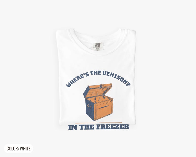 White Comfort Colors t-shirt printed with the words Where'S The Venison? In The Freezer and a drawing of a vintage metal chest freezer with a pair of paper-wrapped butcher packages resting on top, folded flat lay