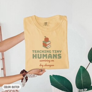 Butter Comfort Colors t-shirt printed with the words Teaching Tiny Humans Surviving On Dry Shampoo and a drawing of a small neat stack of three closed hardcover books with PLAIN blank solid-colour spines an, folded flat lay