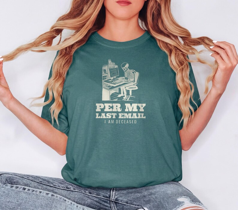 Blue Spruce Comfort Colors t-shirt printed with the words Per My Last Email I Am Deceased and a drawing of a skeleton sitting at an office desk typing on a computer keyboard, worn by a woman