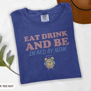 True Navy Comfort Colors t-shirt printed with the words Eat Drink And Be In Bed By Nine and a drawing of a retro brass wind-up alarm clock with its bells wrapped in fuzzy winter earmuffs, folded flat lay