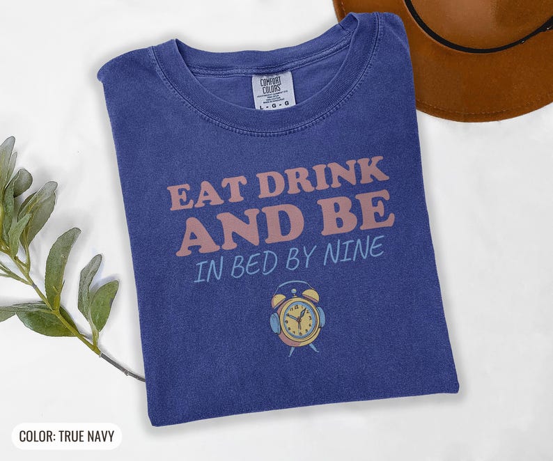True Navy Comfort Colors t-shirt printed with the words Eat Drink And Be In Bed By Nine and a drawing of a retro brass wind-up alarm clock with its bells wrapped in fuzzy winter earmuffs, folded flat lay
