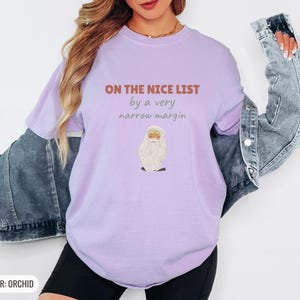 Orchid Comfort Colors t-shirt printed with the words On The Nice List By A Very Narrow Margin and a drawing of a red santa hat beside a candy cane, worn by a woman