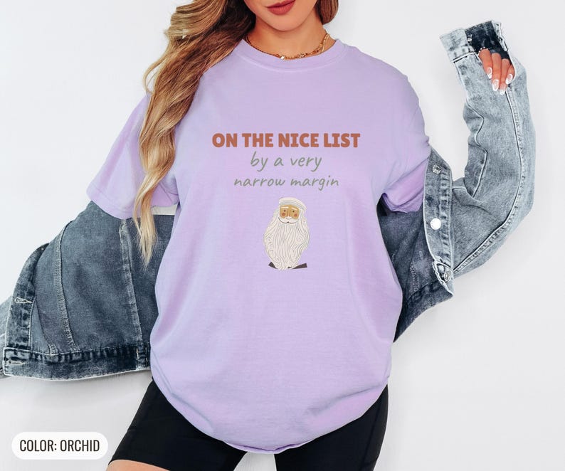 Orchid Comfort Colors t-shirt printed with the words On The Nice List By A Very Narrow Margin and a drawing of a red santa hat beside a candy cane, worn by a woman