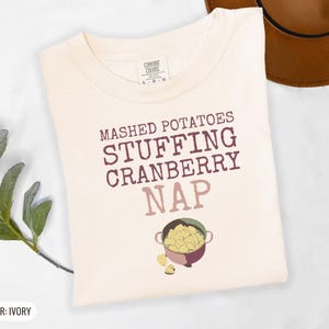 Ivory Comfort Colors t-shirt printed with the words Mashed Potatoes Stuffing Cranberry Nap and a drawing of a heavy enamel-coated cast iron Dutch oven filled with creamy mashed potatoes, steam lines, folded flat lay