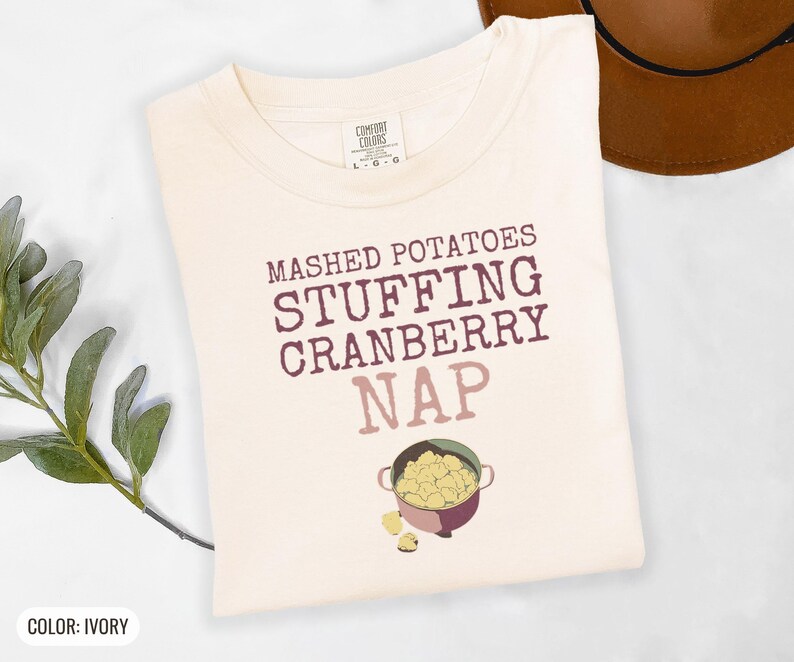 Ivory Comfort Colors t-shirt printed with the words Mashed Potatoes Stuffing Cranberry Nap and a drawing of a heavy enamel-coated cast iron Dutch oven filled with creamy mashed potatoes, steam lines, folded flat lay