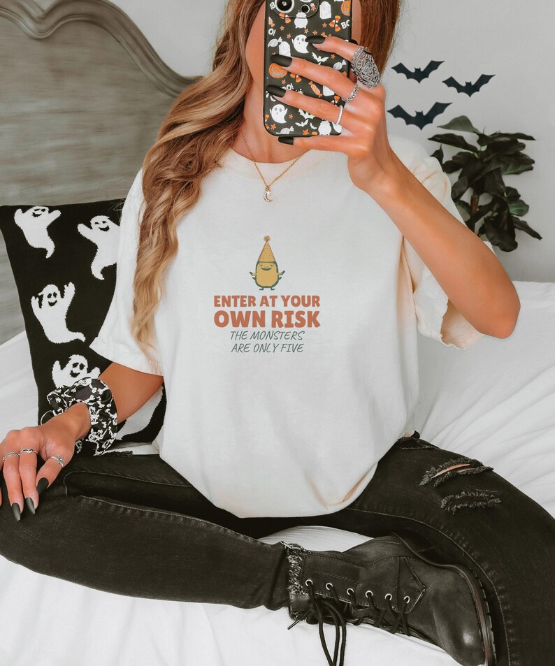Ivory Comfort Colors t-shirt printed with the words Enter At Your Own Risk The Monsters Are Only Five and a drawing of a cute, smiling little green monster wearing a tiny paper party hat and holding a crayon, worn by a woman