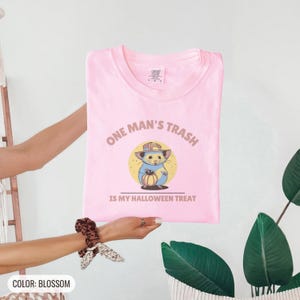 Blossom Comfort Colors t-shirt printed with the words One Man'S Trash Is My Halloween Treat and a drawing of a scruffy opossum wearing a tiny plastic orange pumpkin trick-or-treat bucket on its head, folded flat lay