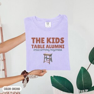 Orchid Comfort Colors t-shirt printed with the words The Kids Table Alumni Still Sitting Together and a drawing of a low wooden folding stool, folded flat lay
