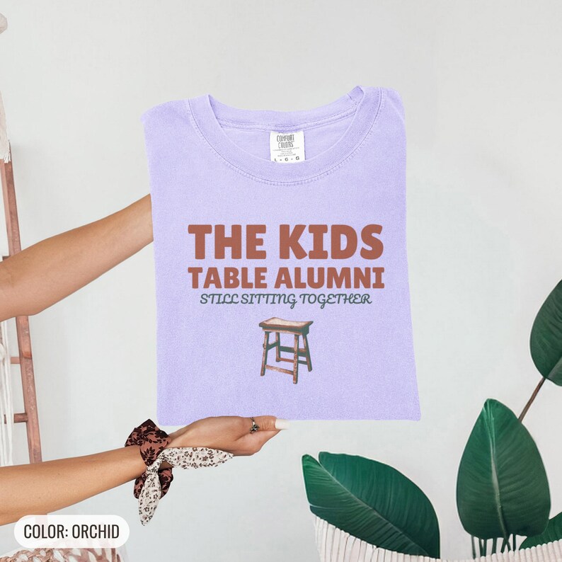 Orchid Comfort Colors t-shirt printed with the words The Kids Table Alumni Still Sitting Together and a drawing of a low wooden folding stool, folded flat lay