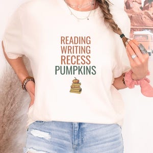 Ivory Comfort Colors t-shirt printed with the words Reading Writing Recess Pumpkins and a drawing of a stack of three vintage schoolbooks with a small orange pumpkin sitting on top, worn by a woman