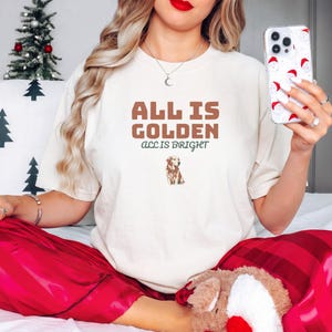 Ivory Comfort Colors t-shirt printed with the words All Is Golden All Is Bright and a drawing of a golden retriever wearing a festive red holiday collar with brass bells, worn by a woman