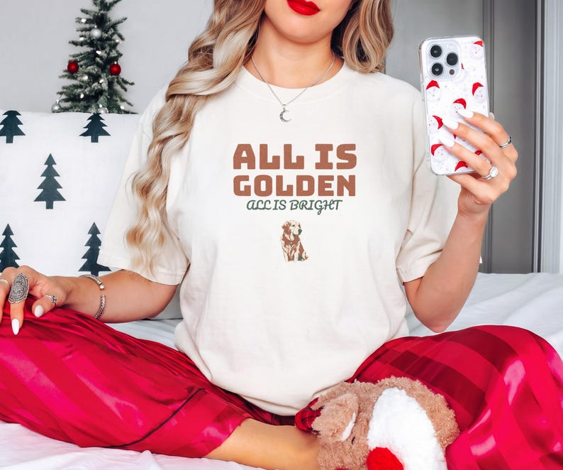 Ivory Comfort Colors t-shirt printed with the words All Is Golden All Is Bright and a drawing of a golden retriever wearing a festive red holiday collar with brass bells, worn by a woman