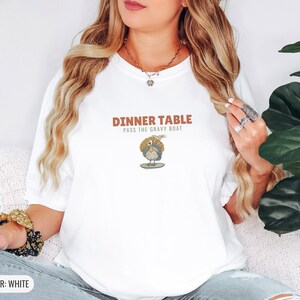 White Comfort Colors t-shirt printed with the words Dinner Table Pass The Gravy Boat and a drawing of a golden roast turkey on a platter, worn by a woman