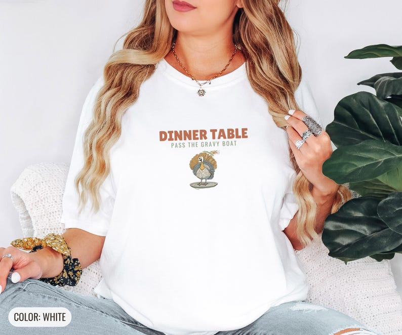 White Comfort Colors t-shirt printed with the words Dinner Table Pass The Gravy Boat and a drawing of a golden roast turkey on a platter, worn by a woman