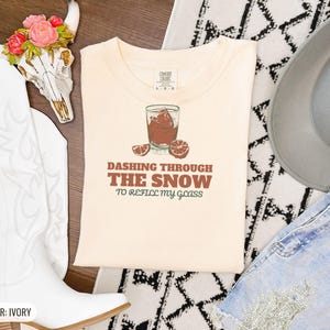 Ivory Comfort Colors t-shirt printed with the words Dashing Through The Snow To Refill My Glass and a drawing of a heavy crystal whiskey tumbler with two floating ice cubes beside a sprig of pine needles, folded flat lay
